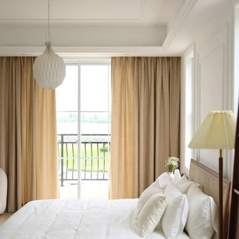 Chambre lumineuse et élégante avec lit double, rideaux beiges et lampe de chevet. Fenêtre donnant sur un balcon avec vue sur la nature. Conception harmonieuse pour une ambiance relaxante et moderne.