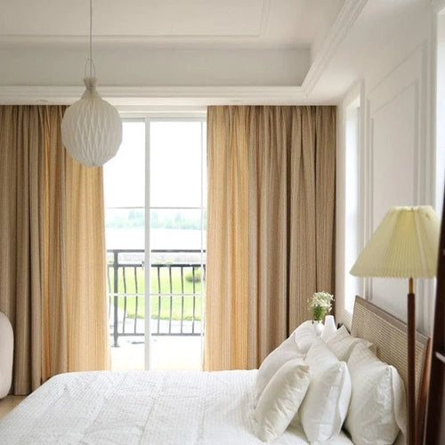 Chambre lumineuse et élégante avec lit double, rideaux beiges et lampe de chevet. Fenêtre donnant sur un balcon avec vue sur la nature. Conception harmonieuse pour une ambiance relaxante et moderne.