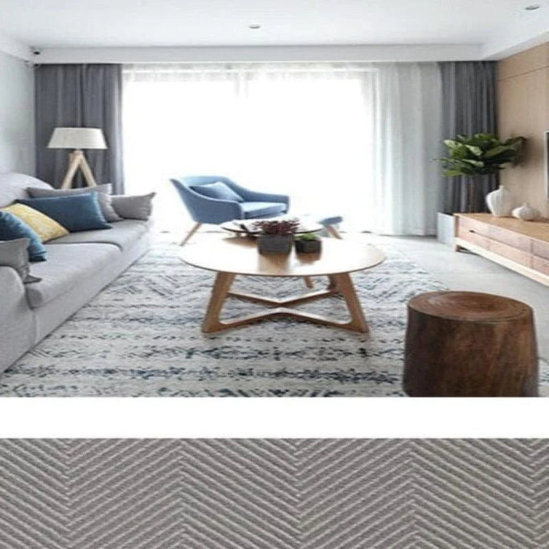 Salon moderne et lumineux avec canapé gris, fauteuil bleu et table basse en bois, accentués par un tapis au motif géométrique. Ambiance chaleureuse grâce aux accessoires décoratifs et à la luminosité naturelle provenant de la grande baie vitrée. Idéal pour une décoration intérieure contemporaine et épurée.
