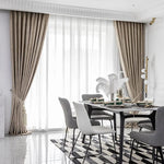 Salle à manger élégante avec rideaux longs et ondulés, baignée de lumière grâce à une grande porte-fenêtre. Table moderne entourée de chaises grises et blanches sur un tapis géométrique monochrome. Décoration raffinée avec objets déco contemporains et éclairage subtil mettant en valeur un intérieur chic et sophistiqué. Idéal pour créer une ambiance accueillante et lumineuse.
