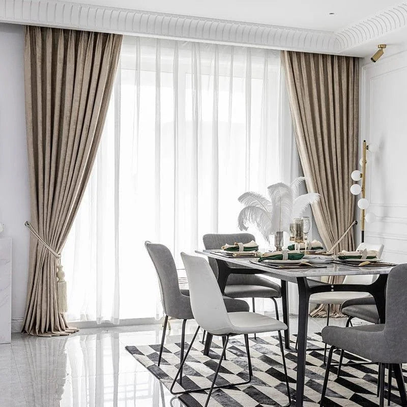 Salle à manger élégante avec rideaux longs et ondulés, baignée de lumière grâce à une grande porte-fenêtre. Table moderne entourée de chaises grises et blanches sur un tapis géométrique monochrome. Décoration raffinée avec objets déco contemporains et éclairage subtil mettant en valeur un intérieur chic et sophistiqué. Idéal pour créer une ambiance accueillante et lumineuse.