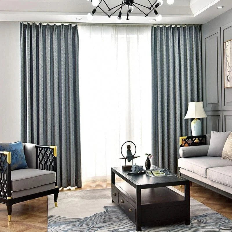 Salon moderne et élégant avec canapé gris, fauteuil design et table basse noire. Les rideaux bleus encadrent une grande fenêtre, apportant une touche de sophistication à la pièce. La décoration raffinée inclut une lampe de table et des accessoires contemporains, créant une ambiance chaleureuse et accueillante. Ce salon est idéal pour un intérieur harmonieux et tendance.