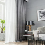 Un élégant salon moderne avec des rideaux gris et blancs, un lampadaire design sur une table noire, et un cadre artistique. La pièce offre une ambiance cosy et contemporaine avec un canapé confortable, des bougies décoratives, et une plante verte apportant une touche de nature. L'éclairage naturel entre par la fenêtre, créant une atmosphère chaleureuse et accueillante.