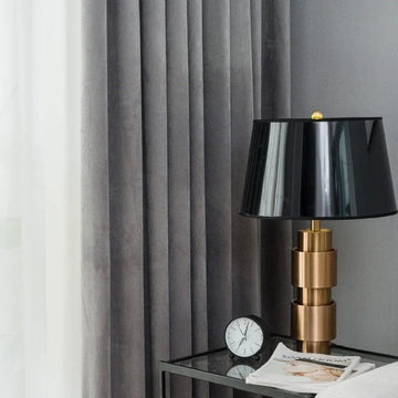Lampe design moderne avec abat-jour noir, posée sur une table en verre à côté d'une horloge, créant une ambiance élégante dans un intérieur contemporain avec rideaux gris.