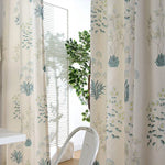 Rideaux décoratifs avec motif floral vert et bleu, suspendus dans une pièce lumineuse. Meuble de bureau blanc et chaise blanche moderne visibles. Ambiance naturelle et apaisante, idéale pour un intérieur élégant.