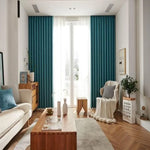 Salon moderne et élégant avec un canapé beige et un coussin bleu, rideaux turquoise, table basse en bois et fauteuil enveloppé d’un plaid. Décoration intérieure lumineuse et soignée, plancher en bois, plantes vertes et accents déco harmonieux. Large fenêtre laissant entrer une lumière naturelle abondante, créant une ambiance chaleureuse et accueillante. Idéal pour inspiration décoration ou aménagement intérieur.