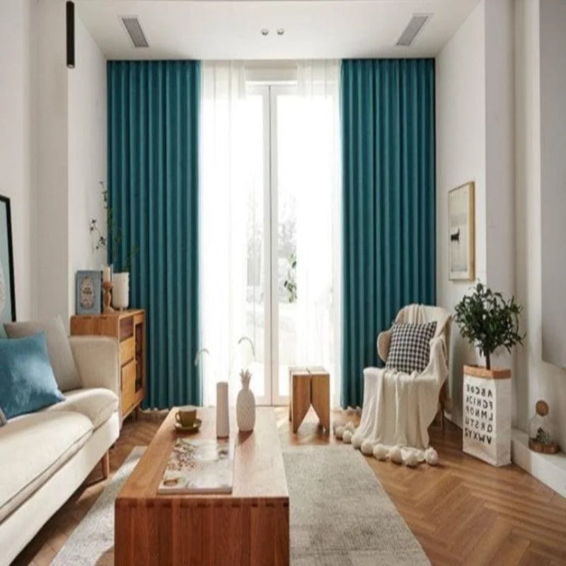 Salon moderne et élégant avec un canapé beige et un coussin bleu, rideaux turquoise, table basse en bois et fauteuil enveloppé d’un plaid. Décoration intérieure lumineuse et soignée, plancher en bois, plantes vertes et accents déco harmonieux. Large fenêtre laissant entrer une lumière naturelle abondante, créant une ambiance chaleureuse et accueillante. Idéal pour inspiration décoration ou aménagement intérieur.