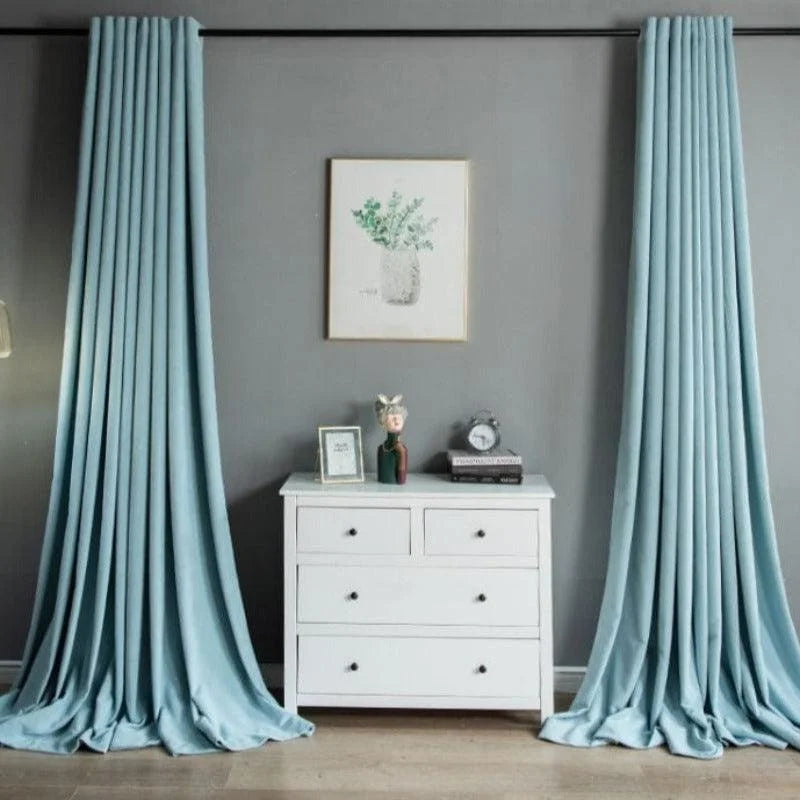 Rideaux bleus élégants pour salon moderne, encadrant une commode blanche sur un mur gris. Décoration murale artistique et accessoires ajoutent une touche de sophistication à l'intérieur. Idéal pour embellir votre pièce à vivre avec style et raffinement.