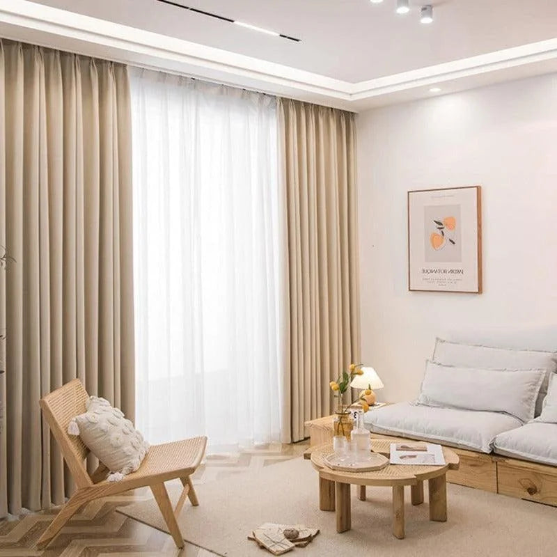 Salon moderne et épuré avec rideaux beige, apportant une ambiance chaleureuse et élégante. Le design minimaliste inclut un canapé confortable avec coussins clairs, une chaise en bois invitante et une table basse assortie. Un tableau ajoute une touche artistique à l'ensemble, créant une atmosphère accueillante et harmonieuse. Idéal pour ceux qui recherchent une décoration intérieure contemporaine et apaisante.