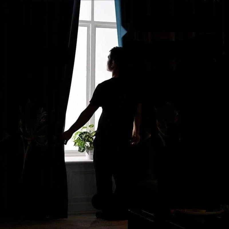 Homme ouvrant des rideaux noirs dans une pièce sombre, révélant une fenêtre lumineuse et une plante verte. Ambiance de tranquillité et d'intimité, parfaite pour la gestion de la lumière naturelle dans votre intérieur. Rideaux élégants pour un style contemporain et raffiné.
