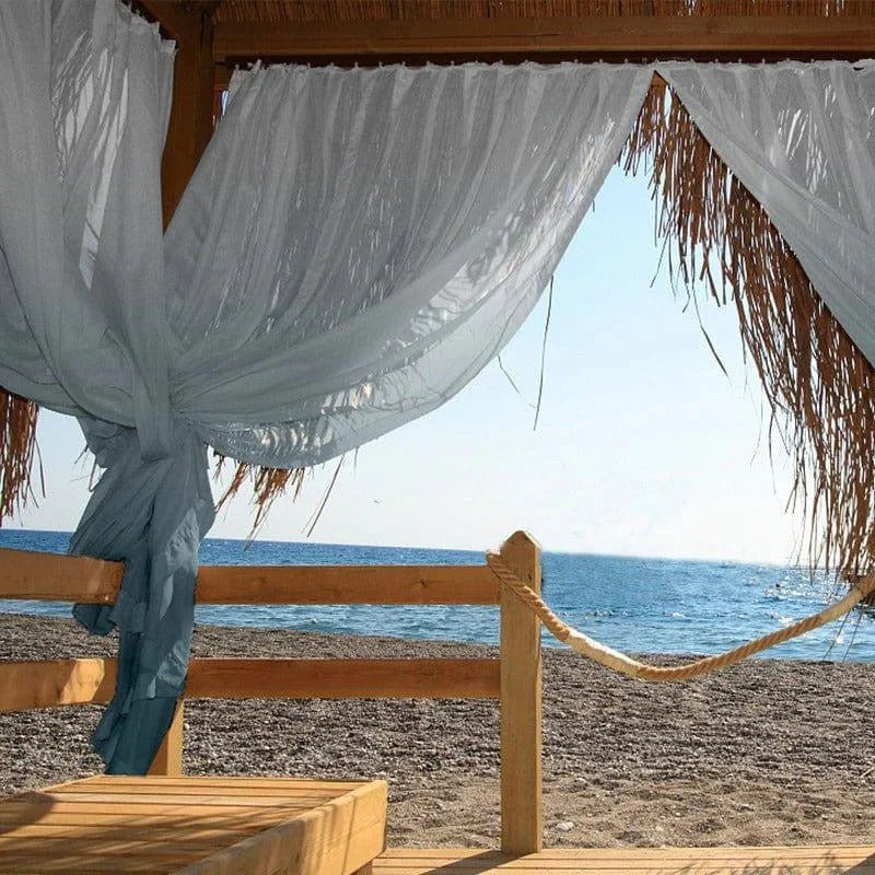 Vue dégagée sur la plage depuis une terrasse ombragée avec rideaux en bord de mer. Cadre idéal pour des moments de détente face à l'océan. Ambiance estivale et relaxation assurée.