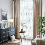 Salon élégant avec rideaux beige et voilages imprimés de motifs floraux, offrant une ambiance lumineuse et paisible. Décoration moderne avec commode noire, lampe design, cadre photo, et plantes vertes. Atmosphère apaisante idéale pour créer un espace de détente harmonieux, parfait pour votre intérieur.