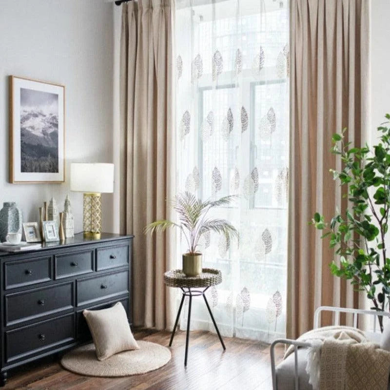 Salon élégant avec rideaux beige et voilages imprimés de motifs floraux, offrant une ambiance lumineuse et paisible. Décoration moderne avec commode noire, lampe design, cadre photo, et plantes vertes. Atmosphère apaisante idéale pour créer un espace de détente harmonieux, parfait pour votre intérieur.