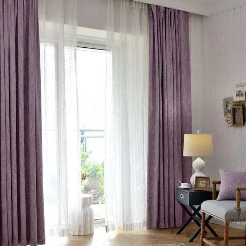 Rideaux opaques violets pour salon, créant une ambiance élégante et moderne. Également visibles, rideaux translucides blancs laissant filtrer la lumière naturelle. Les rideaux encadrent une fenêtre ouverte sur un balcon verdoyant. Le décor inclut une petite table noire avec une lampe élégante, un coussin assorti et des cadres photo muraux. Idéal pour un intérieur contemporain et chaleureux.