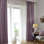 Rideaux opaques violets pour salon, créant une ambiance élégante et moderne. Également visibles, rideaux translucides blancs laissant filtrer la lumière naturelle. Les rideaux encadrent une fenêtre ouverte sur un balcon verdoyant. Le décor inclut une petite table noire avec une lampe élégante, un coussin assorti et des cadres photo muraux. Idéal pour un intérieur contemporain et chaleureux.