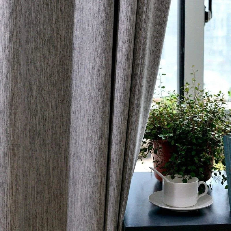 Rideau moderne gris dans un salon lumineux, avec une plante d'intérieur et une tasse sur une table à proximité d'une fenêtre en arrière-plan. Idéal pour créer une ambiance chaleureuse et intime. Parfait pour un intérieur contemporain et élégant.