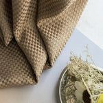 Tissu décoratif avec motif tissé brun, présenté sur une surface claire avec un arrangement floral minimaliste. Parfait pour ajouter une touche de sophistication et de naturel à votre intérieur. Design élégant et intemporel avec des accents naturels.