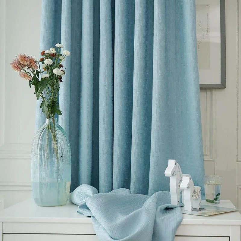 Rideaux élégants de couleur bleu clair dans un intérieur moderne, accompagnés d'un vase décoratif avec des fleurs fraîches sur une table blanche, parfaits pour ajouter une touche de sérénité à votre salon ou chambre.