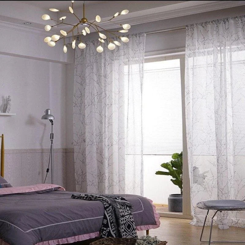 Chambre moderne et élégante avec un lustre design en forme de branche, rideaux translucides imprimés de motifs subtils, et lit soigneusement aménagé avec une couverture grise. Lampadaire ajustable et plante verte ajoutent une touche de confort naturel. Idéal pour une ambiance relaxante et contemporaine.