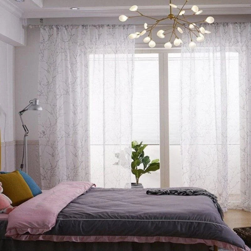 Chambre élégante avec lit double et couette grise, rehaussée par des coussins colorés. Les rideaux aux motifs subtils filtrent la lumière naturelle, créant une atmosphère paisible. Suspension contemporaine et plante d'intérieur ajoutant une touche moderne et chaleureuse. Convient pour une décoration intérieure chic et accueillante.