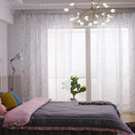 Chambre élégante avec lit double et couette grise, rehaussée par des coussins colorés. Les rideaux aux motifs subtils filtrent la lumière naturelle, créant une atmosphère paisible. Suspension contemporaine et plante d'intérieur ajoutant une touche moderne et chaleureuse. Convient pour une décoration intérieure chic et accueillante.