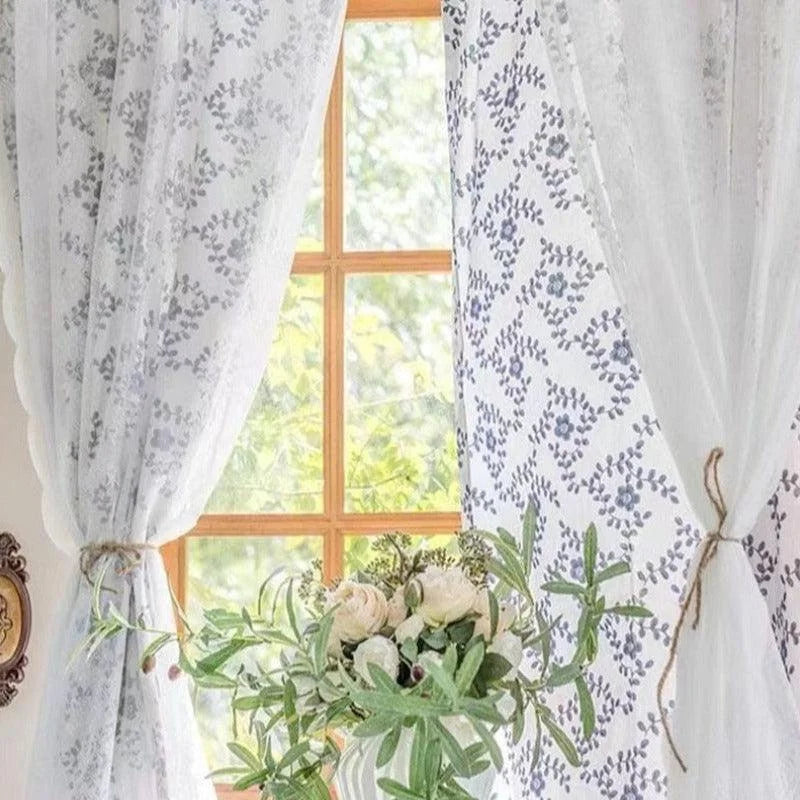 Rideaux élégants à motifs floraux devant une fenêtre en bois, offrant une ambiance lumineuse et naturelle. Une composition florale blanche et verte ajoute une touche de fraîcheur et de décoration intérieure à cet espace chaleureux. Styles de rideaux tendance pour embellir votre maison.