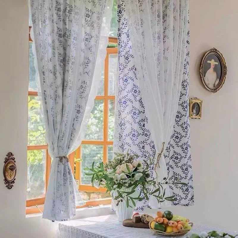 Rideaux blancs avec motif floral, décorant une fenêtre lumineuse dans une ambiance intérieure chaleureuse. Une table est agrémentée d'un bouquet de fleurs et d'un assortiment de fruits, créant une atmosphère accueillante et naturelle.  Accents de décoration murale ajoutent une touche d'élégance au cadre environnant.