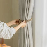 Personne attachant une élégante cordelette autour d'un rideau beige pour un style raffiné. Décoration intérieure moderne et fonctionnelle pour un salon lumineux et accueillant. Solution pratique pour gérer la luminosité tout en ajoutant une touche décorative. Idéal pour améliorer l'ambiance de votre espace de vie.