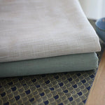 Deux tissus élégants empilés, l'un beige et l'autre vert, posés sur une nappe aux motifs décoratifs dorés et noirs, parfaits pour la décoration d'intérieur et l'aménagement d'espaces modernes et sophistiqués.