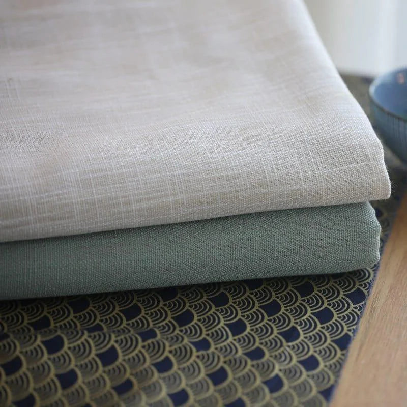 Deux tissus élégants empilés, l'un beige et l'autre vert, posés sur une nappe aux motifs décoratifs dorés et noirs, parfaits pour la décoration d'intérieur et l'aménagement d'espaces modernes et sophistiqués.