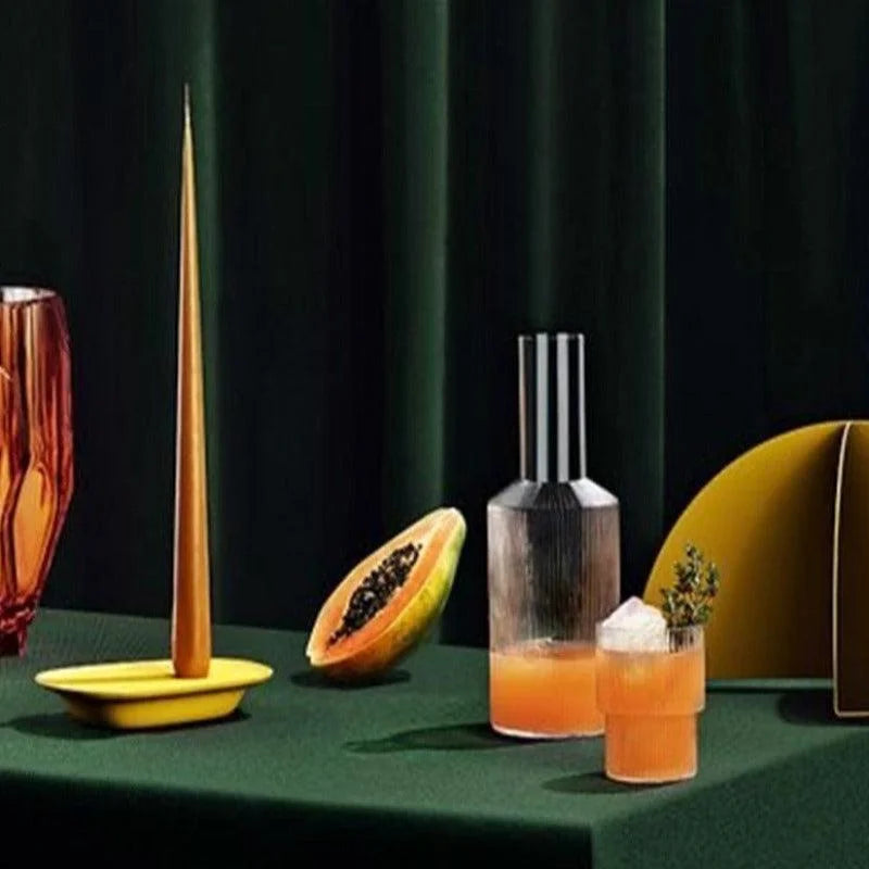 Composition d'un centre de table moderne avec une élégante carafe et un verre contenant une boisson orange. Présentés sur une nappe verte, des éléments décoratifs incluent une bougie effilée jaune, un récipient sculpté et une moitié de papaye. Ce décor élégant et coloré est idéal pour inspirer une atmosphère conviviale et chaleureuse lors d'un dîner ou d'une réception.