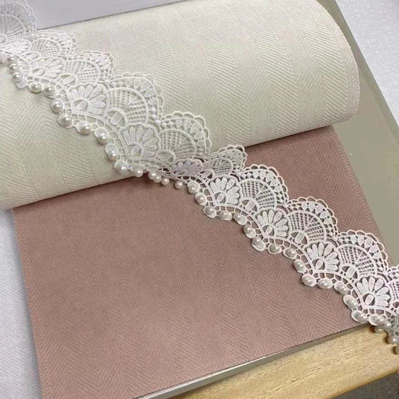 Dentelle délicate blanche ornée de perles élégantes sur un fond pastel, idéale pour la décoration intérieure et les projets de couture créatifs. Parfait pour ajouter une touche raffinée à vos créations artisanales.