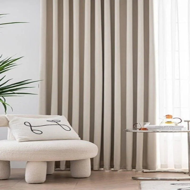 Fauteuil moderne beige avec coussin décoratif devant un rideau élégant, table basse métallique avec théière, intérieur contemporain et lumineux, ambiance sophistiquée et accueillante.