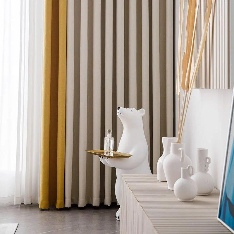 Décoration intérieure moderne avec un ours décoratif portant un plateau doré, placé devant des rideaux élégants en dégradé de jaune et beige. Vases blancs design sur une console contemporaine, complétant un environnement chic et raffiné. Accessoire décoratif unique pour un salon sophistiqué.