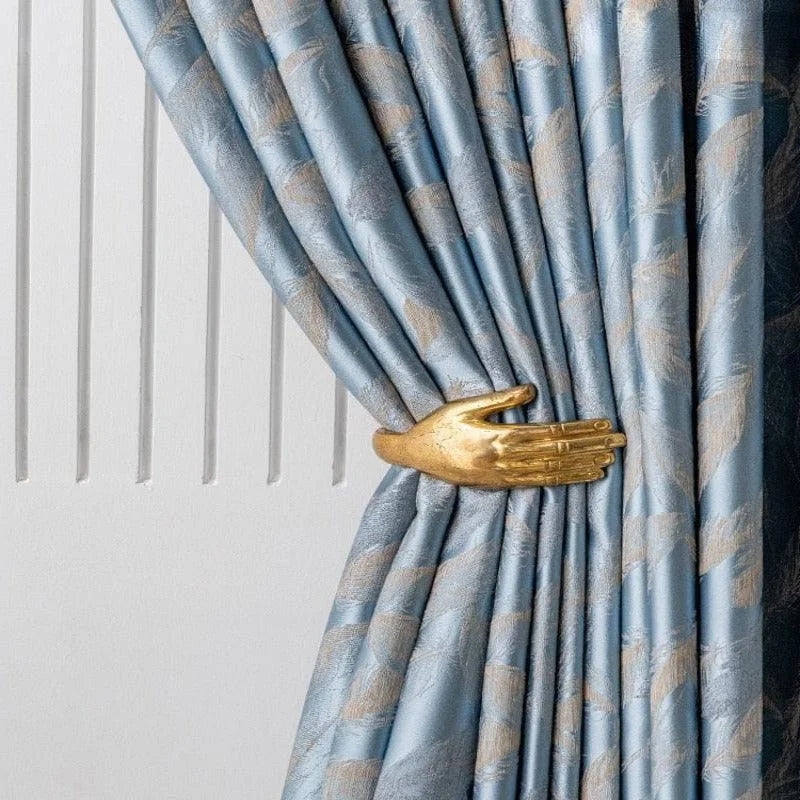 Attache-rideau élégant en forme de main en or, ajoutant une touche artistique et sophistiquée à un rideau bleu à motifs. Accessoire de décoration d'intérieur design pour sublimer votre espace de vie avec originalité.