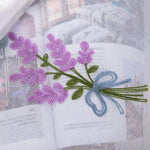 Broderie florale délicate représentant un bouquet de fleurs violettes avec feuilles vertes, ornée d'un nœud bleu. Design élégant parfait pour la décoration de textiles, ajoutant une touche artistique et raffinée à tout projet de couture ou de décoration intérieure. Motif idéal pour personnaliser vêtements ou accessoires avec une esthétique florale charmante et moderne.