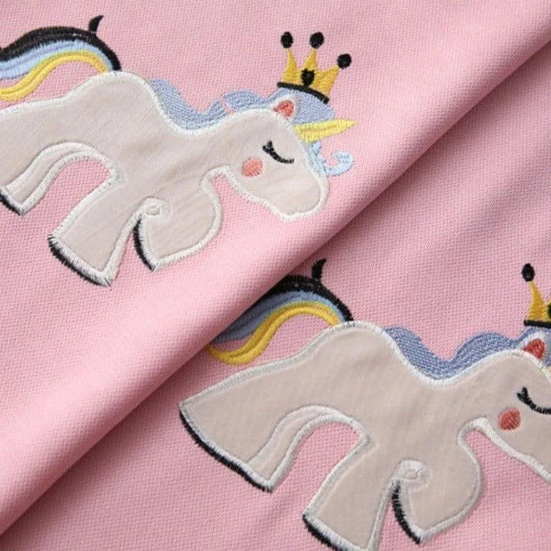 Tissu rose avec motif licorne brodé, couronné d'une couronne dorée, crinière arc-en-ciel, design enfantin, idéal pour décoration ou couture thématique.
