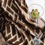 Rideau moderne avec motif géométrique en zigzag marron et beige, idéal pour ajouter une touche élégante à votre intérieur. Accessoire décoratif parfait pour salon ou chambre, associé à un décor naturel comprenant une table en bois et une coupe en verre avec un artichaut décoratif.