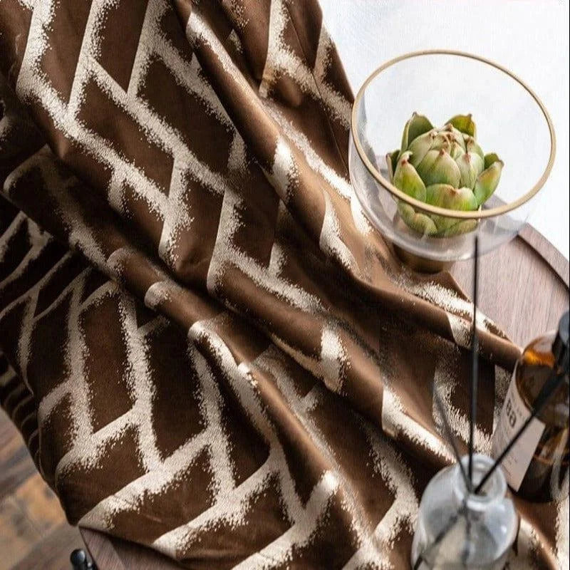 Rideau moderne avec motif géométrique en zigzag marron et beige, idéal pour ajouter une touche élégante à votre intérieur. Accessoire décoratif parfait pour salon ou chambre, associé à un décor naturel comprenant une table en bois et une coupe en verre avec un artichaut décoratif.