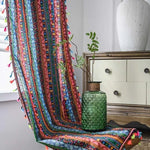 Image de décoration intérieure comprenant un meuble en bois, une nappe multicolore à motifs géométriques avec des franges colorées et un vase décoratif vert en verre texturé. L'ensemble crée une ambiance chaleureuse et artistique, idéale pour un salon moderne. L'éclairage naturel met en valeur les détails de la composition.