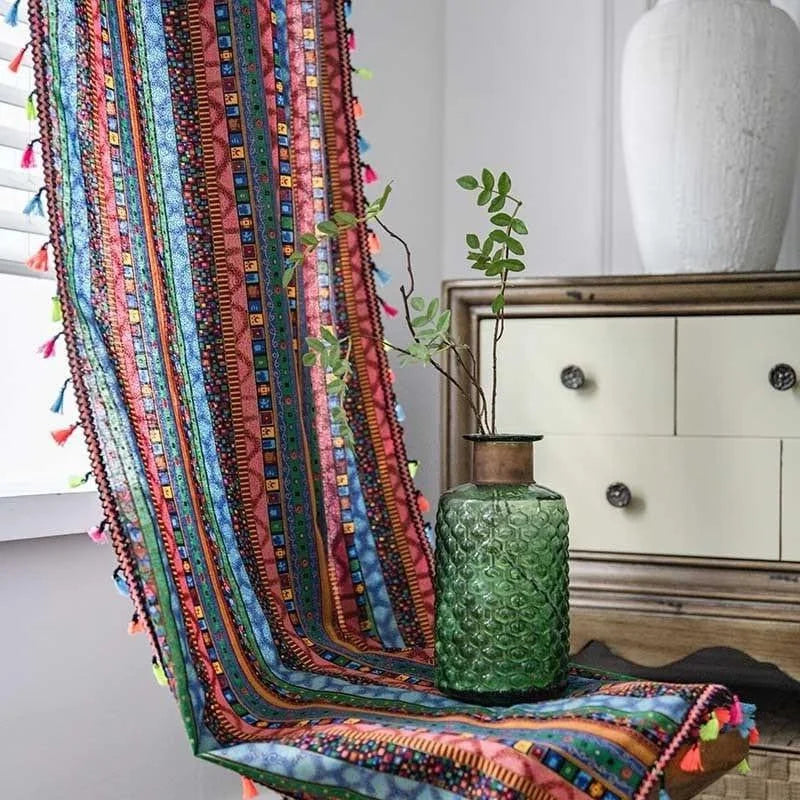 Image de décoration intérieure comprenant un meuble en bois, une nappe multicolore à motifs géométriques avec des franges colorées et un vase décoratif vert en verre texturé. L'ensemble crée une ambiance chaleureuse et artistique, idéale pour un salon moderne. L'éclairage naturel met en valeur les détails de la composition.