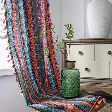 Image de décoration intérieure comprenant un meuble en bois, une nappe multicolore à motifs géométriques avec des franges colorées et un vase décoratif vert en verre texturé. L'ensemble crée une ambiance chaleureuse et artistique, idéale pour un salon moderne. L'éclairage naturel met en valeur les détails de la composition.