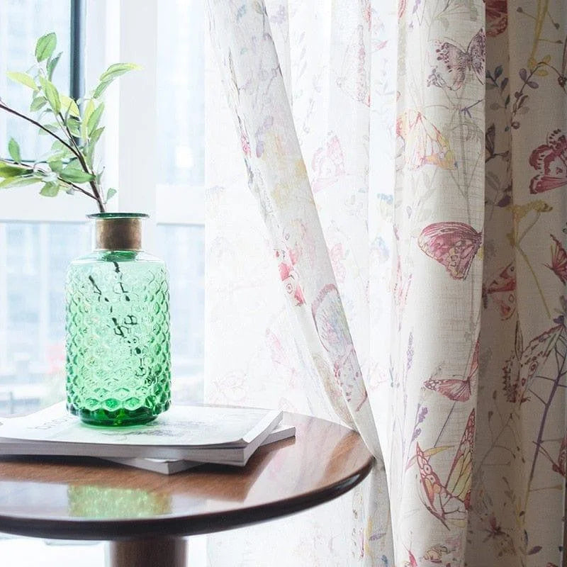 Vase décoratif vert avec motif à relief, posé sur une table en bois, devant une fenêtre habillée de rideaux aux motifs de papillons colorés et de fleurs. Ambiance lumineuse et naturelle idéale pour une décoration de salon moderne.