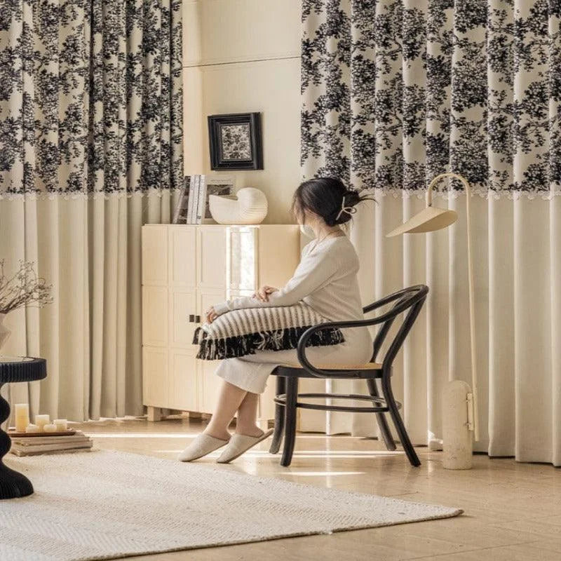 Une femme est assise sur une chaise noire élégante, regardant vers une élégante décoration intérieure. Les rideaux à motifs floraux ajoutent une touche artistique à la pièce, tandis qu'un tapis clair au sol complète l'ambiance douce et moderne de l'espace de vie. Un meuble de rangement discret et une lampe design apportent fonctionnalité et style au décor.
