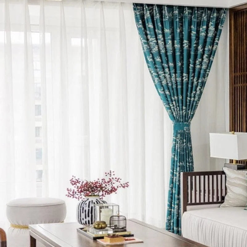 Rideau bleu à motif floral dans un salon moderne, apportant une touche élégante avec des voilages blancs filtrant subtilement la lumière. Décoration d'intérieur raffinée avec table basse en bois, vase décoratif et coussins harmonieux. Ambiance lumineuse et apaisante.
