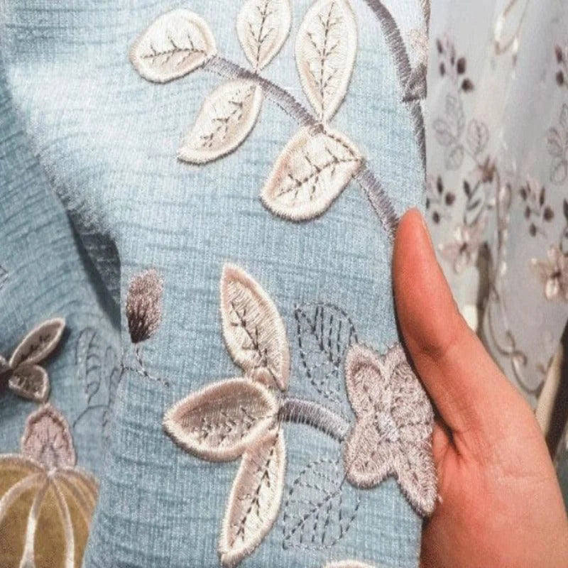 Bel exemple de tissu bleu clair avec motif floral brodé en relief. Ce design représente des feuilles délicatement détaillées, idéal pour rehausser la décoration intérieure. Le tissu est tenu par une main, soulignant la finesse des détails et la qualité de la broderie, parfait pour des rideaux ou des pièces décoratives uniques.