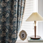 Rideaux élégants aux motifs floraux bleutés dans un intérieur lumineux, avec une lampe à abat-jour plissé sur une table près d'un cadre ovale vintage. Ambiance cosy et raffinée pour une décoration d'intérieur contemporaine.