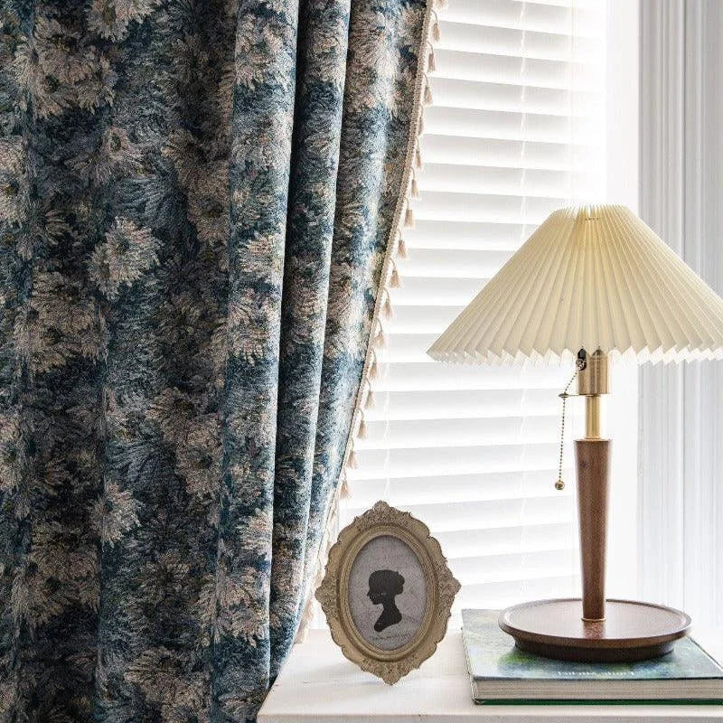 Rideaux élégants aux motifs floraux bleutés dans un intérieur lumineux, avec une lampe à abat-jour plissé sur une table près d'un cadre ovale vintage. Ambiance cosy et raffinée pour une décoration d'intérieur contemporaine.