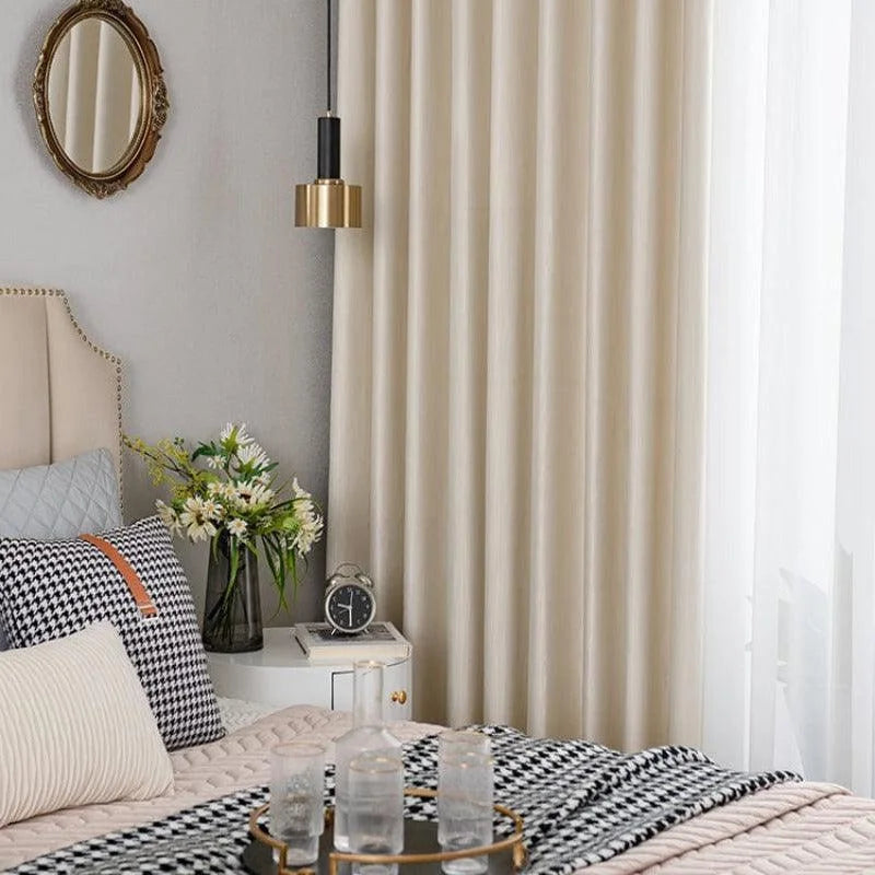 Chambre élégante avec décoration intérieure moderne, comprenant un lit confortable avec des coussins stylisés. Un élégant rideau beige encadre la fenêtre, apportant une touche de sophistication. La chambre est agrémentée d'une table de chevet avec un réveil et un vase de fleurs, offrant une ambiance cosy et agréable.