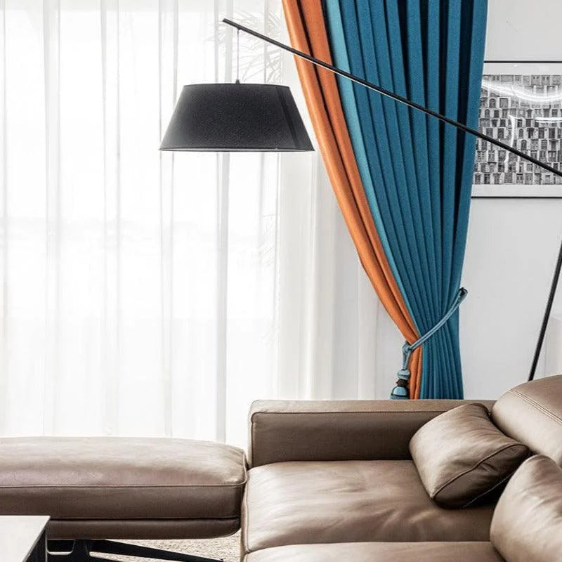 Canapé moderne et élégant avec repose-pieds assorti, idéal pour un salon contemporain. Accompagné de rideaux bicolores bleu et orange apportant une touche de couleur vive à la décoration intérieure. Lampe sur pied au design minimaliste pour un éclairage chic et fonctionnel. Parfait pour créer une ambiance chaleureuse et accueillante.